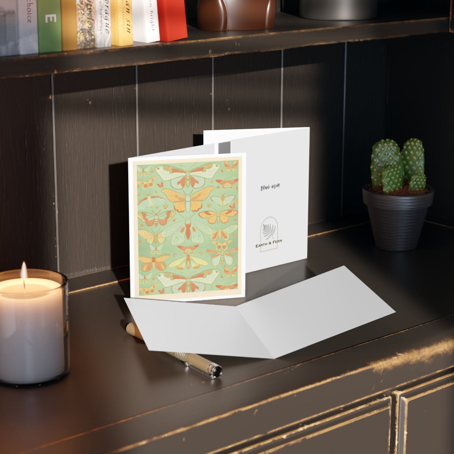 Image shows a greeting card with rows of butterflies in a collection. The butterflies are colored in shades of light mint, yellow and orange. On a medium mint background with a double border of cream and white. Card sits on a desk with a candle , books and a cactus. The inside and back of card are shown. The inside is blank and the back of the card shows the words ”Blank inside” and the Earth and Fern logo below.