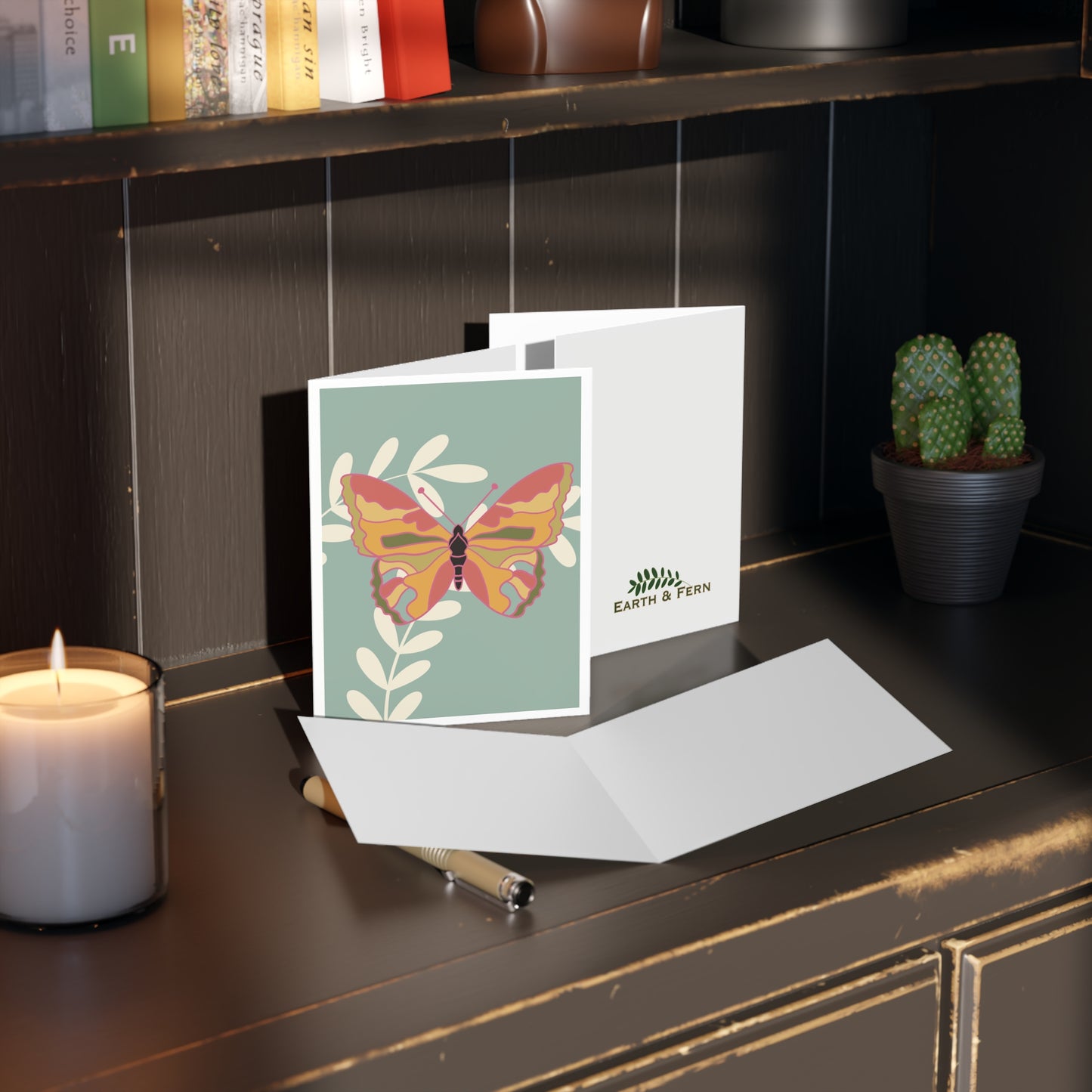Greeting card - Image shows a yellow, orange, and pink butterfly sitting on cream leaves on a light blue background with white border. Cards shows the inside and back of card. The card inside is blank and the back shows the Earth and Fern logo. Card sits on a a desk with a candle, books and a cactus.