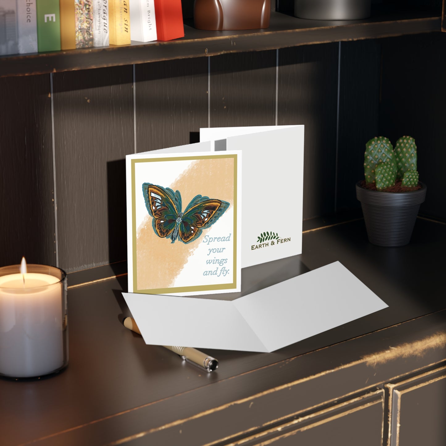 The image shows the front of the greeting card. On it is a Blue, yellow, orange and brown butterfly in flight. Behind is is a streak of faded light orange in its flight path. The background is white with a sage and white double border. The words “Spread your wings and fly” are written on the card. The card sits on a desk and shows the back and inside of the card also. The inside is blank and the back shows the Earth & fern logo with the words “ Blank inside” above it.