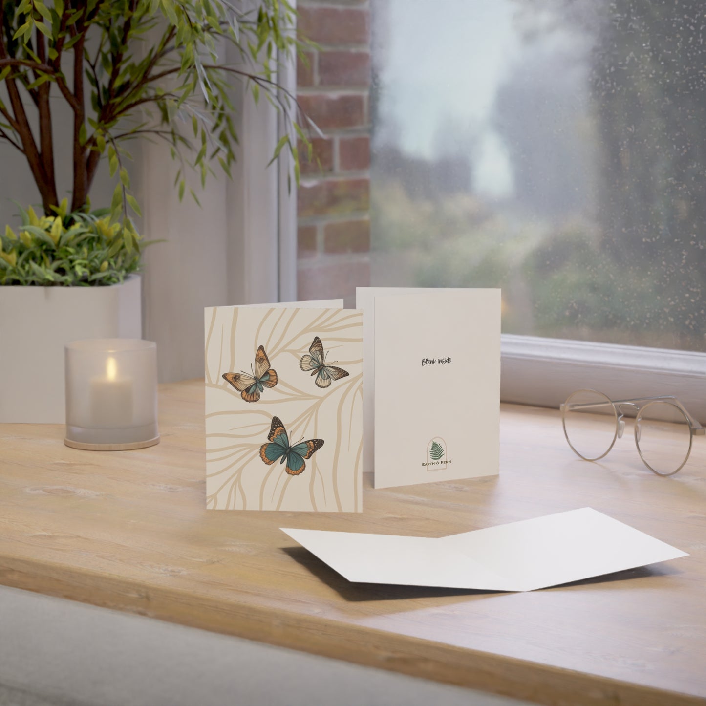 Image shows the front of the greeting card. The Background is cream color with light tan branches covering the whole card. Sitting on the branches are three butterflies of varying size. All in the colors of blue, cream, brown and light orange. The card is sitting on a desk along side a candle, plant and by a window. The card shows the back and inside of card. The inside is blank and the back says “ Blank Inside”. Also it shows the Earth and Fern logo.