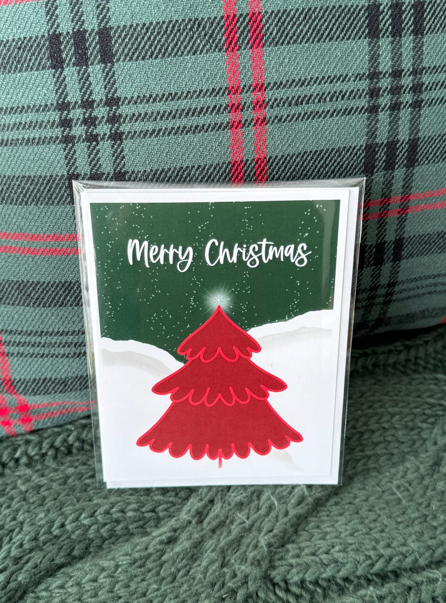Charming greeting card featuring a red Christmas tree adorned with decorations, set against a snowy landscape and a night sky with light snowflakes falling, with the words 'Merry Christmas' elegantly displayed near the top. Card is blank inside and has Earth and Fern logo on the back.