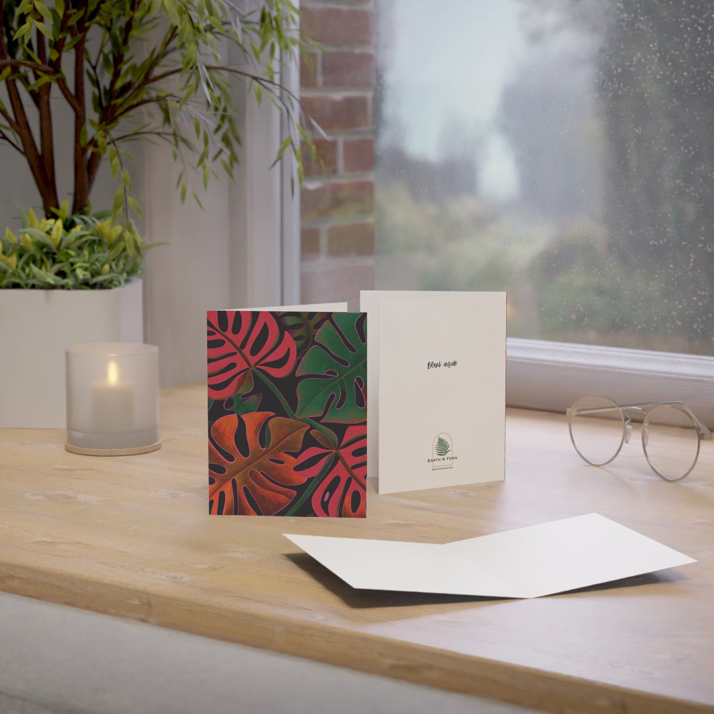 Mockup of Holiday Monstera Greeting Card set showing the front, blank inside, and back. The front features a vibrant tropical Monstera leaf design with holiday colors. The inside is blank for a personalized message. The back showcases the Earth & Fern logo with the words 'Blank Inside.