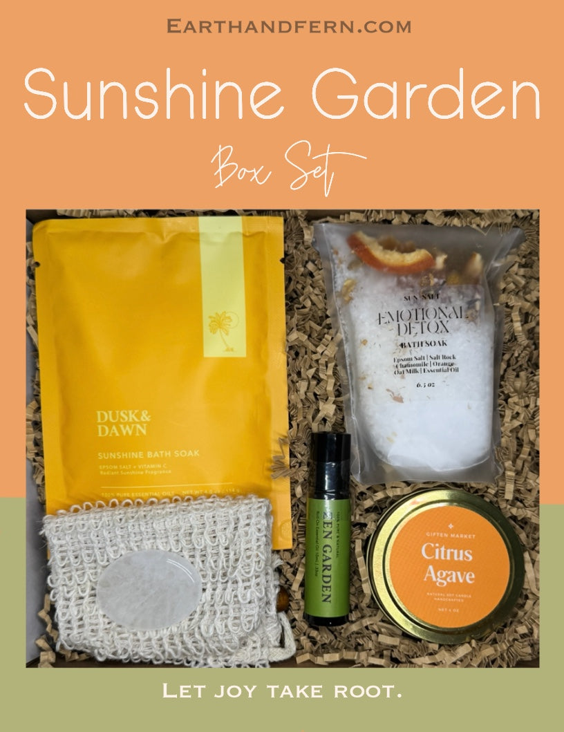 Bright and cheerful wellness gift box called Sunshine Garden, featuring a citrus agave soy candle, citrus peel bath soak, sunshine epsom salt soak, essential oil roll-on, and a clear quartz crystal, arranged in a vibrant yellow, orange, and melon green color theme.