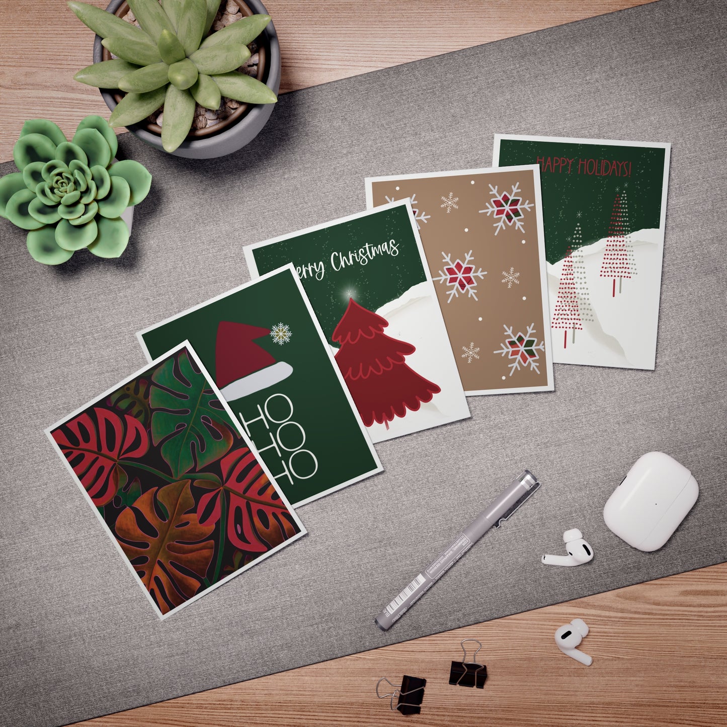 Mockup of a set of five mixed greeting cards displaying the front designs, showing a monstera plant, Santa hat with 'Ho, Ho, Ho,' night sky with a red tree, tan card with snowflakes and plaid, and night sky with skinny pencil Christmas trees. Blank Inside