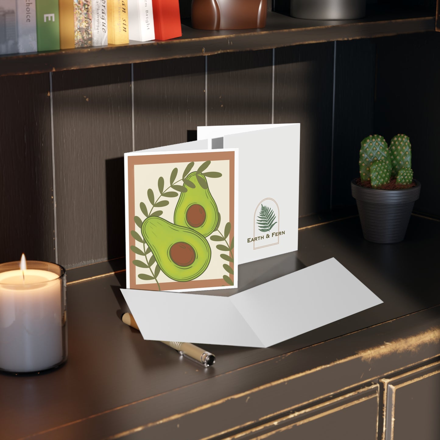 Mockup of greeting card showing front design with brown border, cream background, and avocados, blank inside, and company logo on the back.
