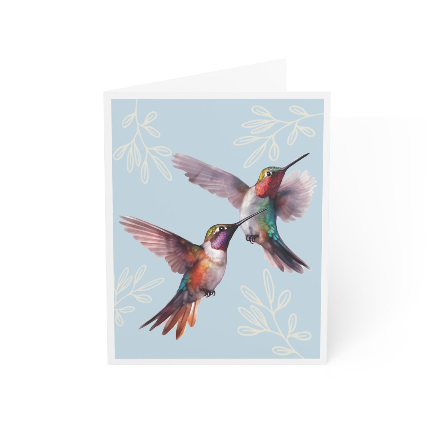 Front of a greeting card with a soft light blue background featuring two vibrant, colorful hummingbirds in flight, drawn in a realistic style. The hummingbirds are surrounded by delicate cream-colored leaves, adding to the natural beauty of the design. The card exudes a peaceful, nature-inspired aesthetic, perfect for sending heartfelt greetings. The inside of the card is blank, ideal for personal messages, and the back includes the text 'Blank inside' along with the company logo