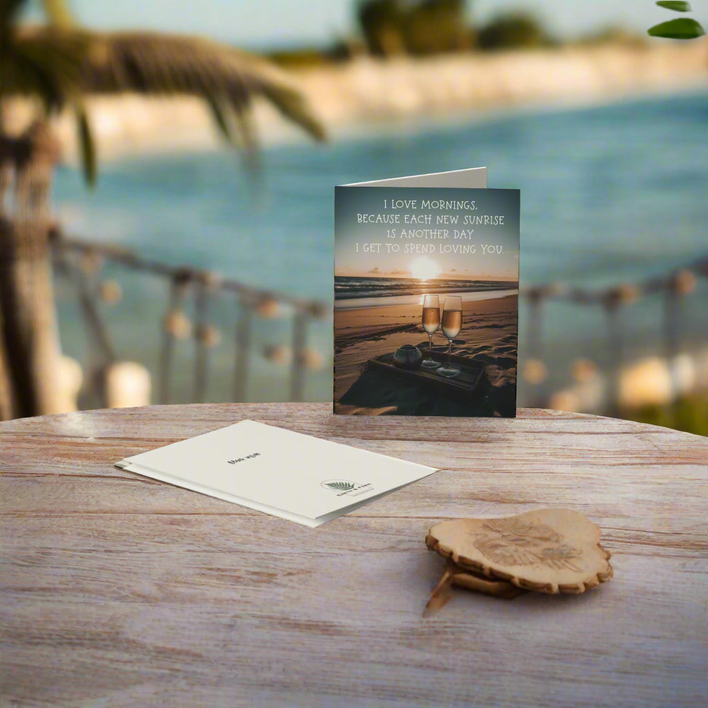 A romantic greeting card featuring a sunrise, two glasses of champagne on a beach blanket, and the message "I love mornings, because each new sunrise is another day I get to spend loving you.