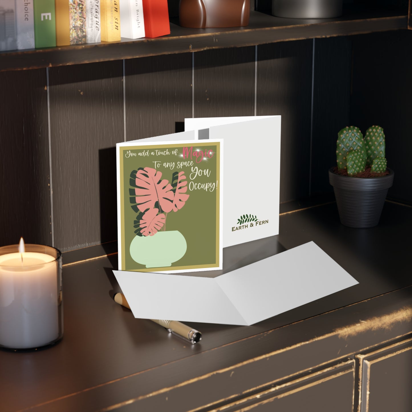 Front of a sage green greeting card with a white border, featuring a mint-colored pot holding a unique pink Monstera plant. The card includes the phrase 'You add a touch of magic to every space you occupy' written in pink, adding a heartfelt message. The card's blank inside is perfect for a personalized note, with the back displaying the text 'Blank inside' and the company logo.