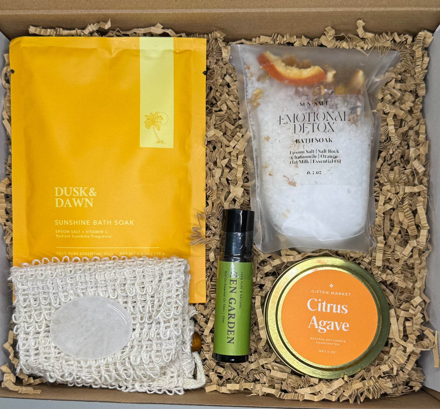 Bright and cheerful wellness gift box called Sunshine Garden, featuring a citrus agave soy candle, citrus peel bath soak, sunshine epsom salt soak, essential oil roll-on, and a clear quartz crystal, arranged in a vibrant yellow, orange, and forest green color theme.