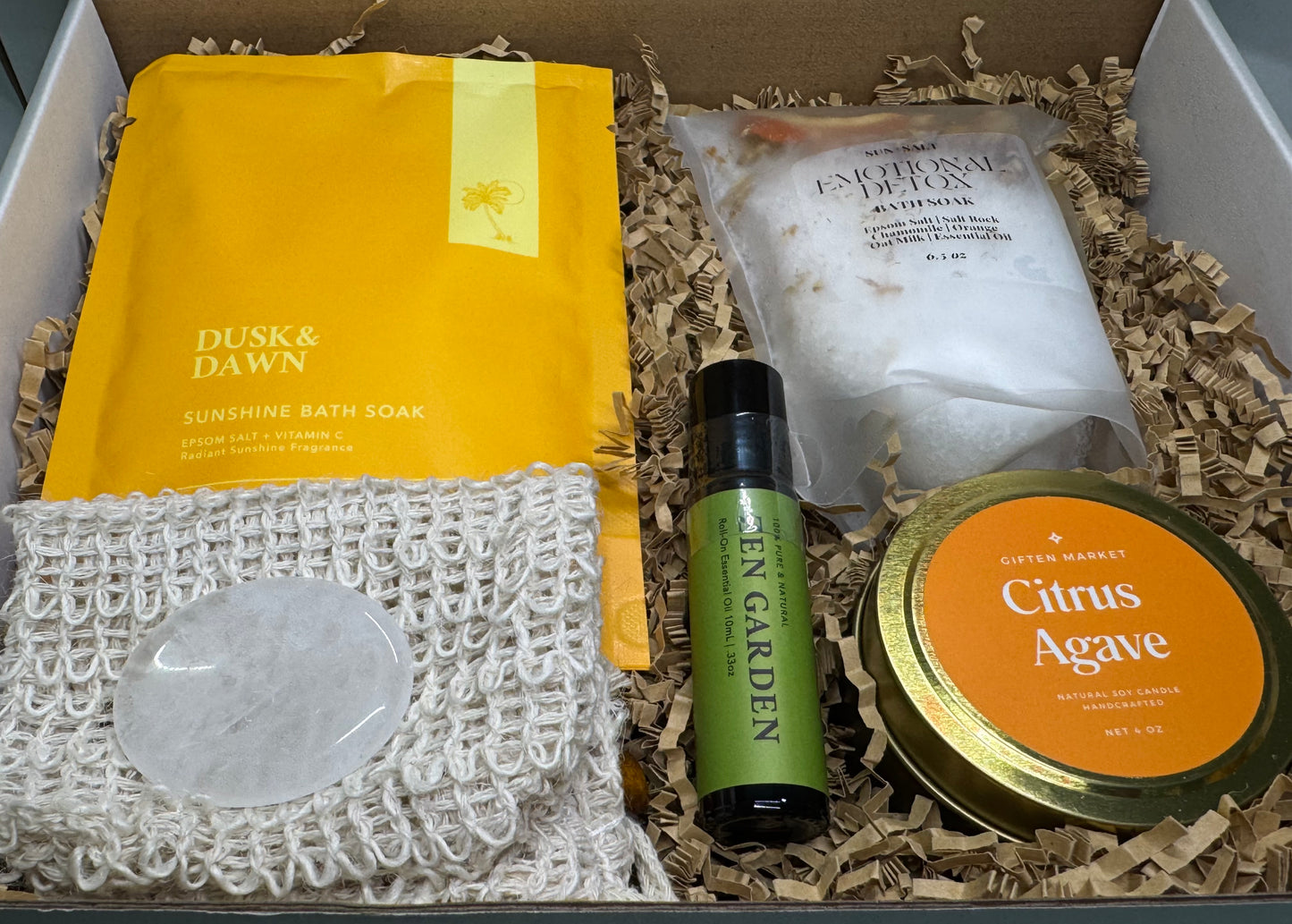Bright and cheerful wellness gift box called Sunshine Garden, featuring a citrus agave soy candle, citrus peel bath soak, sunshine epsom salt soak, essential oil roll-on, and a clear quartz crystal, arranged in a vibrant yellow, orange, and tropical green color theme.