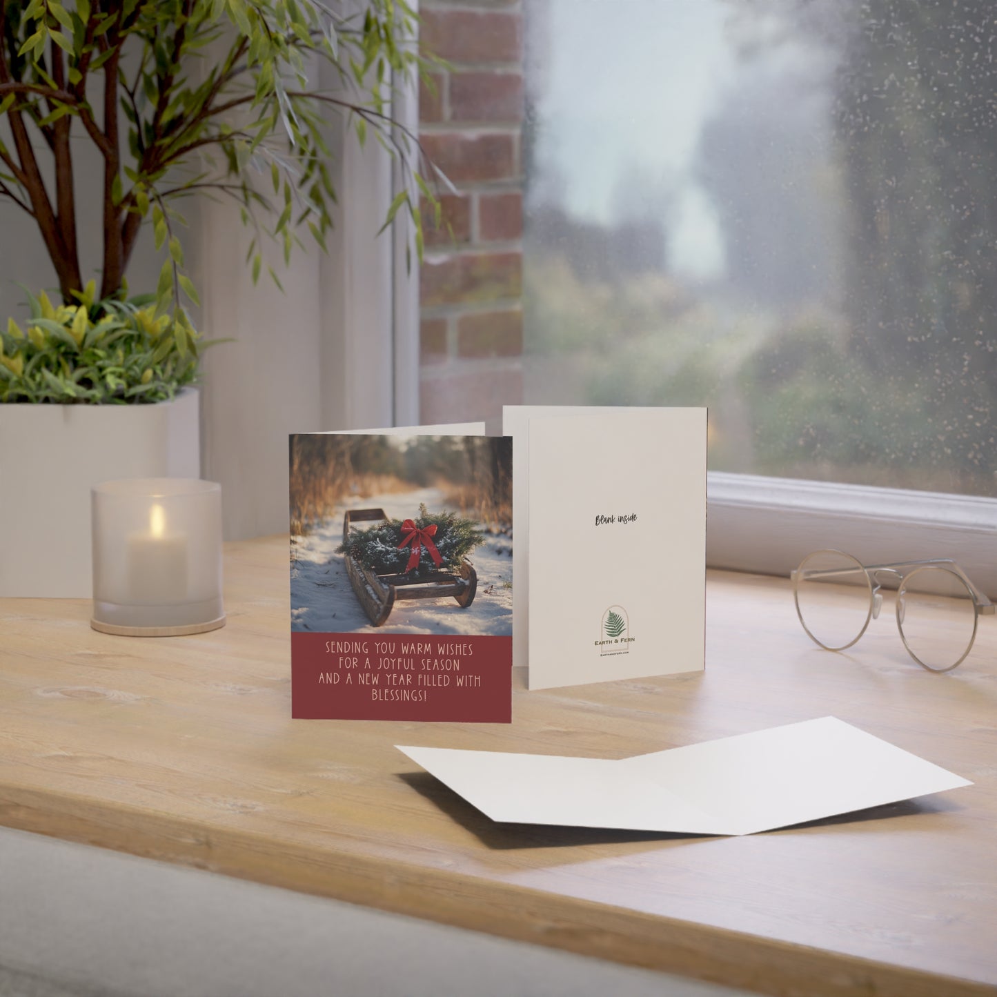 Mockup of holiday greeting card featuring a vintage wooden sled with an evergreen wreath and red bow on a snowy path. The front includes the message: 'Sending you warm wishes for a joyful season and a new year filled with blessings.' The inside is blank for a personalized message. The back features the Earth & Fern logo with the words 'Blank Inside‘.