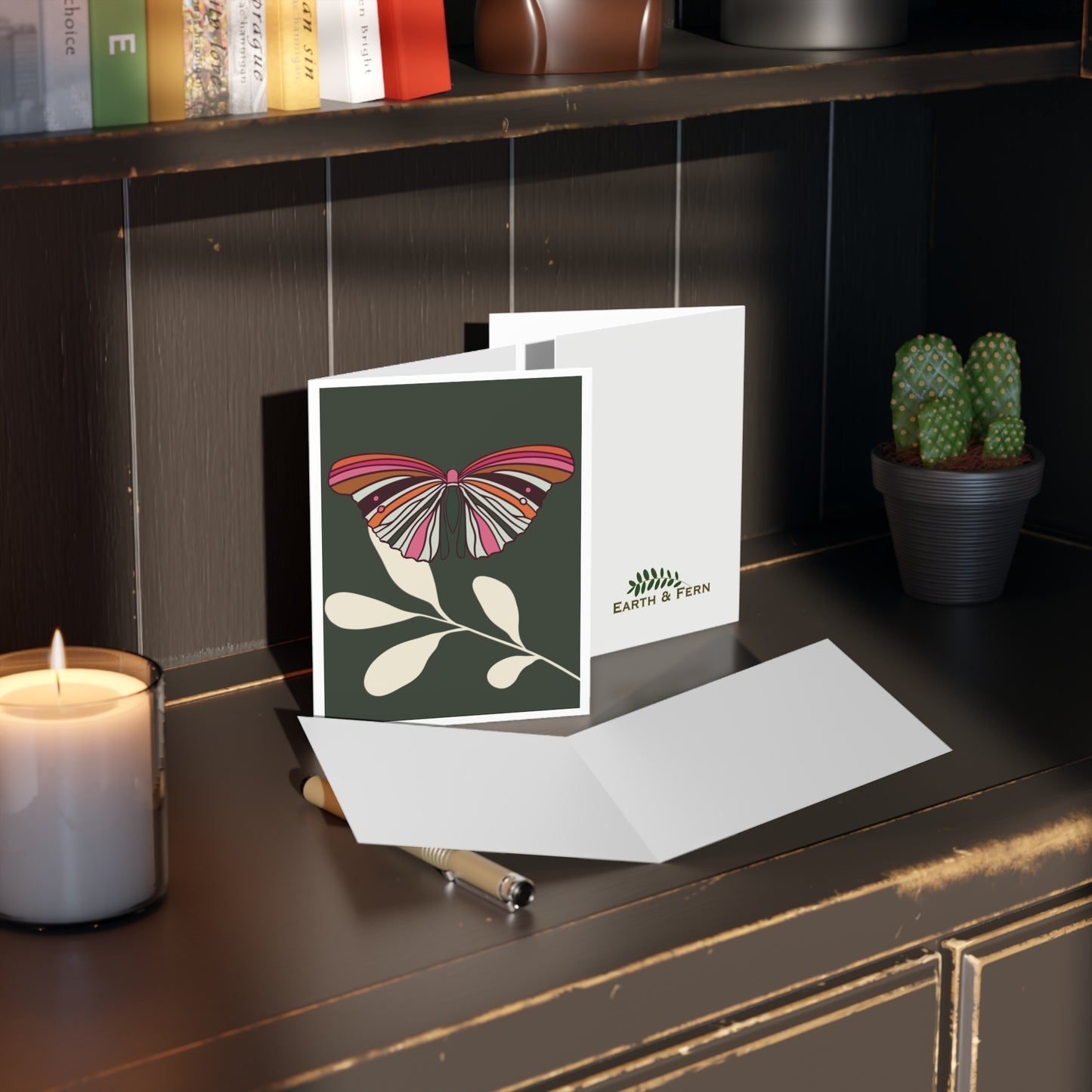 A sophisticated greeting card featuring a vibrant butterfly with wings spread, perched on a cream-colored leaf, set against a deep regency green background, perfect for any occasion. Image shows a Greeting Card with a single colorful butterfly with the colors orange, hot pink, cream and green colors. It sits on a single cream leaf with a forest green background with white border.