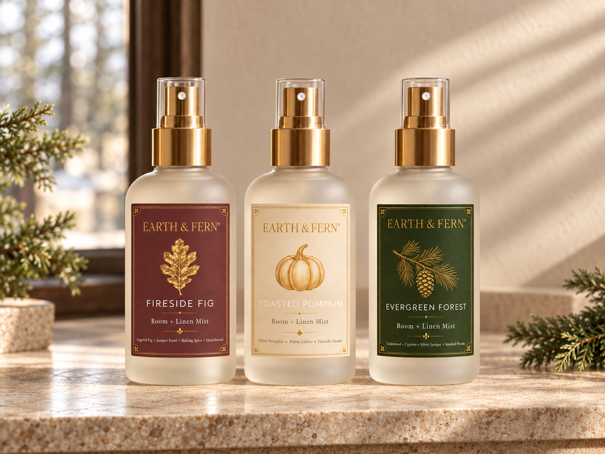 Three Earth & Fern room spray bottles in different scents (Fireside Fig, Toasted Pumpkin, Evergreen Forest) lined up on a stone countertop with warm sunlight and pine branches in the background. Save when you bundle !