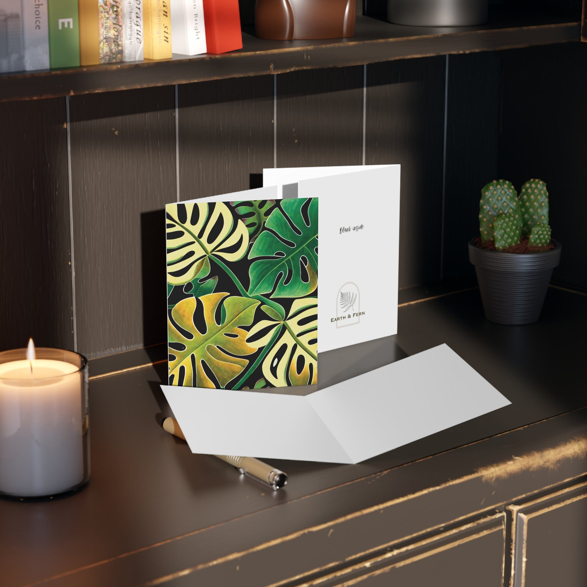 Front of a greeting card featuring a design of four Monstera leaves in various shades of green, arranged in a stylish quad layout. The vibrant green tones of the Monstera leaves create a fresh, tropical look, perfect for plant lovers or nature-themed occasions. The inside of the card is blank for personal messages, with the back displaying the text 'Blank inside' and the company logo.