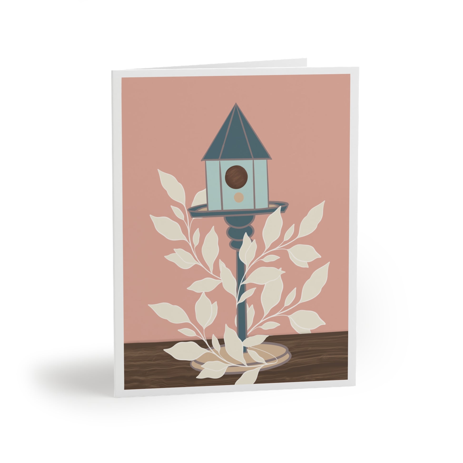 Image shows a greeting card with a blue birdhouse on a stand sits on a wooden table top. Cream leaves wrap around the bird stand, winding their way up. The back ground is salmon coral color with white border.