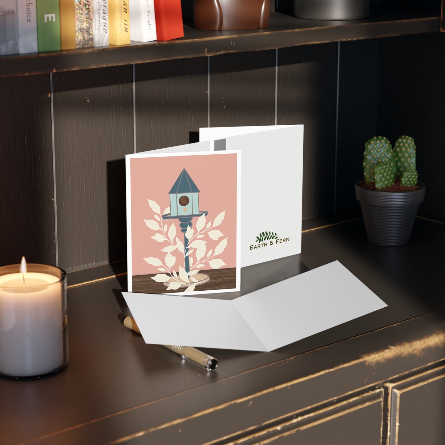 Image shows a greeting card with a blue birdhouse on a stand sits on a wooden table top. Cream leaves wrap around the bird stand, winding their way up. The back ground is salmon coral color with white border. The card sits on a a table with the front and back and inside of card showing. The inside is blank and the back of the card shows the Earth & Fern logo.