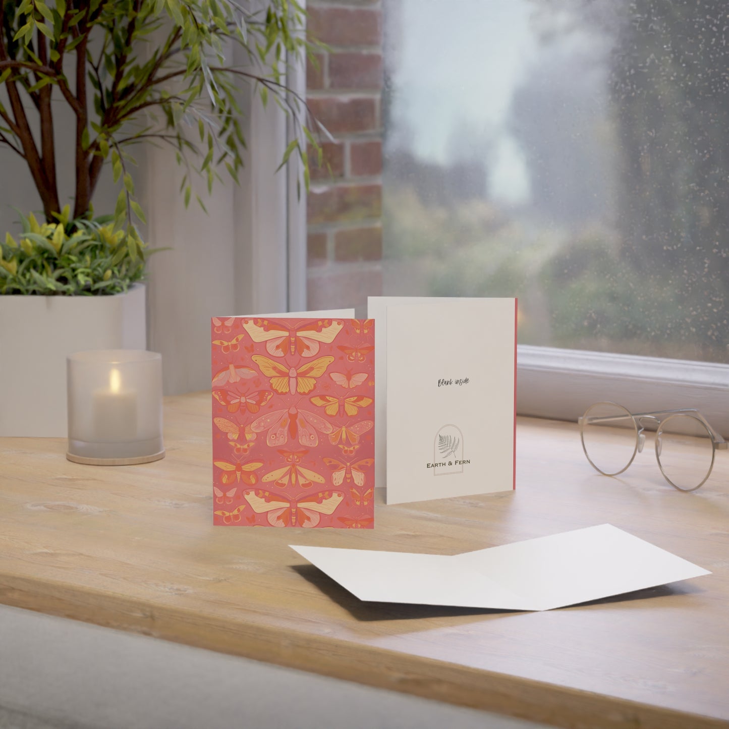 Image shows a greeting card with rows of butterflies in a collection. The butterflies are colored in shades of pink, yellow and orange. On a coral pink background with no border. Card sits on a table by a window with a candle. It shows the back and inside of the card. The inside is blank and the back shows the words  “ Blank inside” and the Earth and Fern logo.