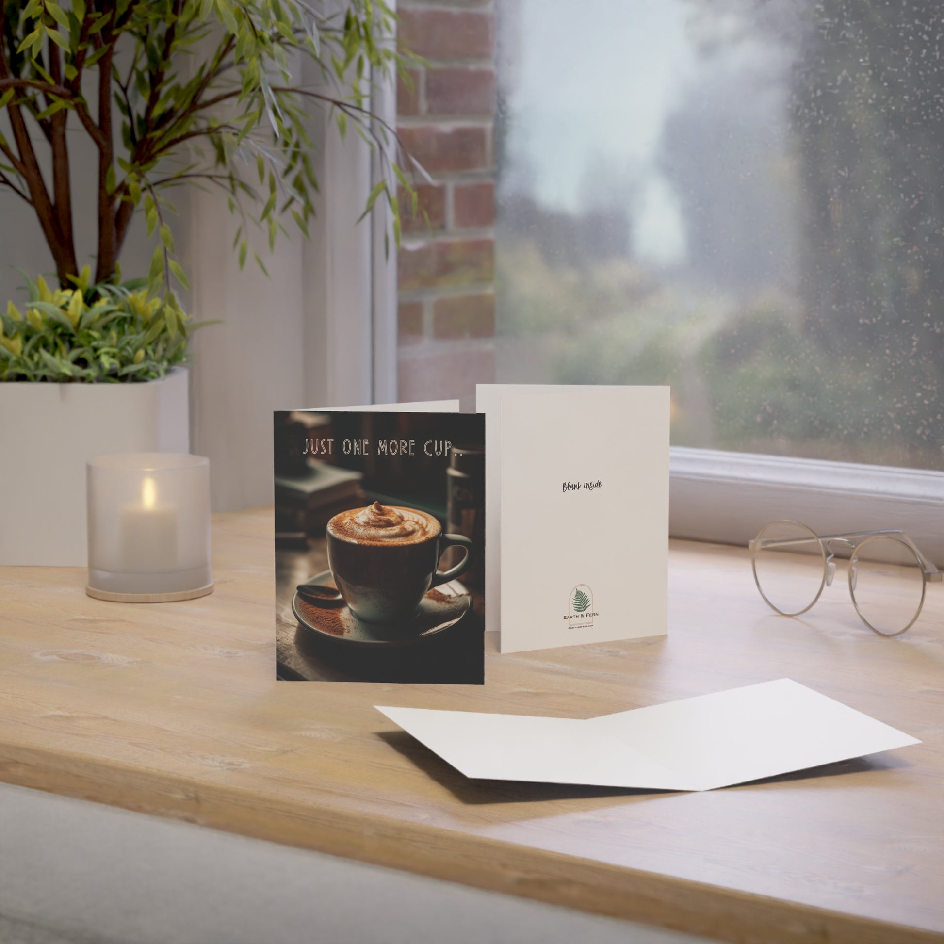 Mockup of a greeting card featuring a cozy coffee shop scene on the front with a steaming cup of coffee and the text  'Just One More Cup..’ above. Inside is blank for personal messages, and the back displays the company logo with the phrase 'Blank inside” above.