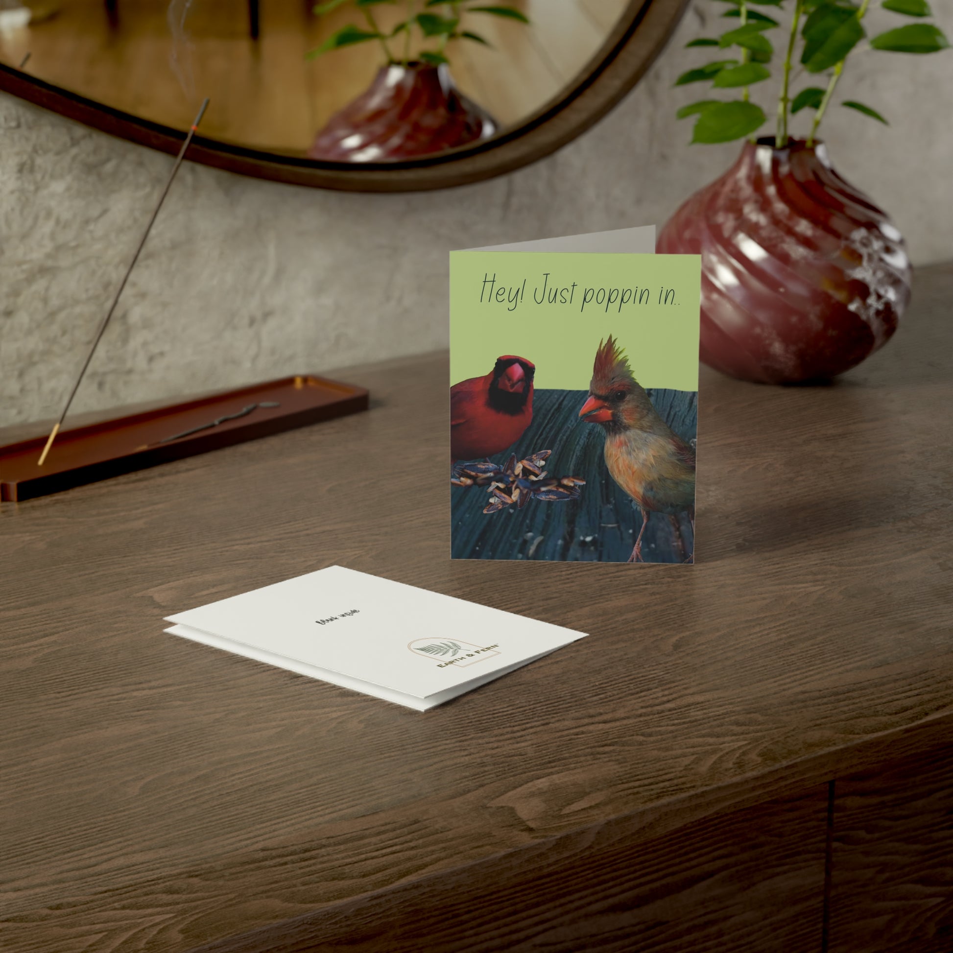 The image shows two cardinal birds peaking at the camera, standing on a piece of wood. The background color of the card is light green with the words  Hey! Just poppin in.. The card sits on a brown table with a vase and a pen. The front and back of card are visible. The back of the card shows the words “blank inside” and the Earth & Fern logo.