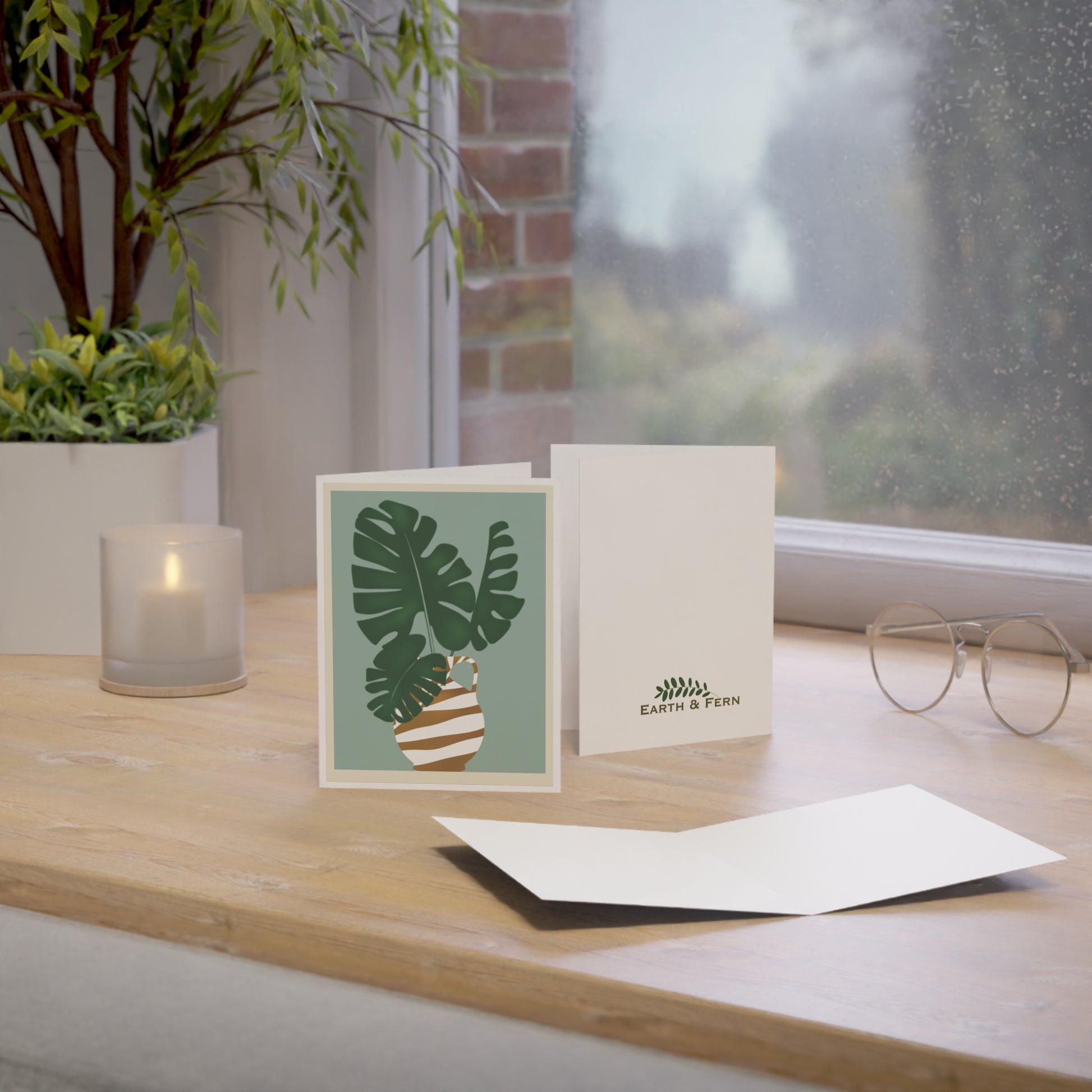 Front of a greeting card featuring a pastel aqua background with a Monstera plant displayed in a gold and white striped vase, with the vase drawn in an ancient Egyptian style. The card is framed by a white border around the perimeter, creating a clean, elegant look. The inside of the card is blank, perfect for personal messages, and the back includes the text 'Blank inside' along with the company logo. Ideal for sending nature-inspired greetings, home decor enthusiasts, or any special occasion.