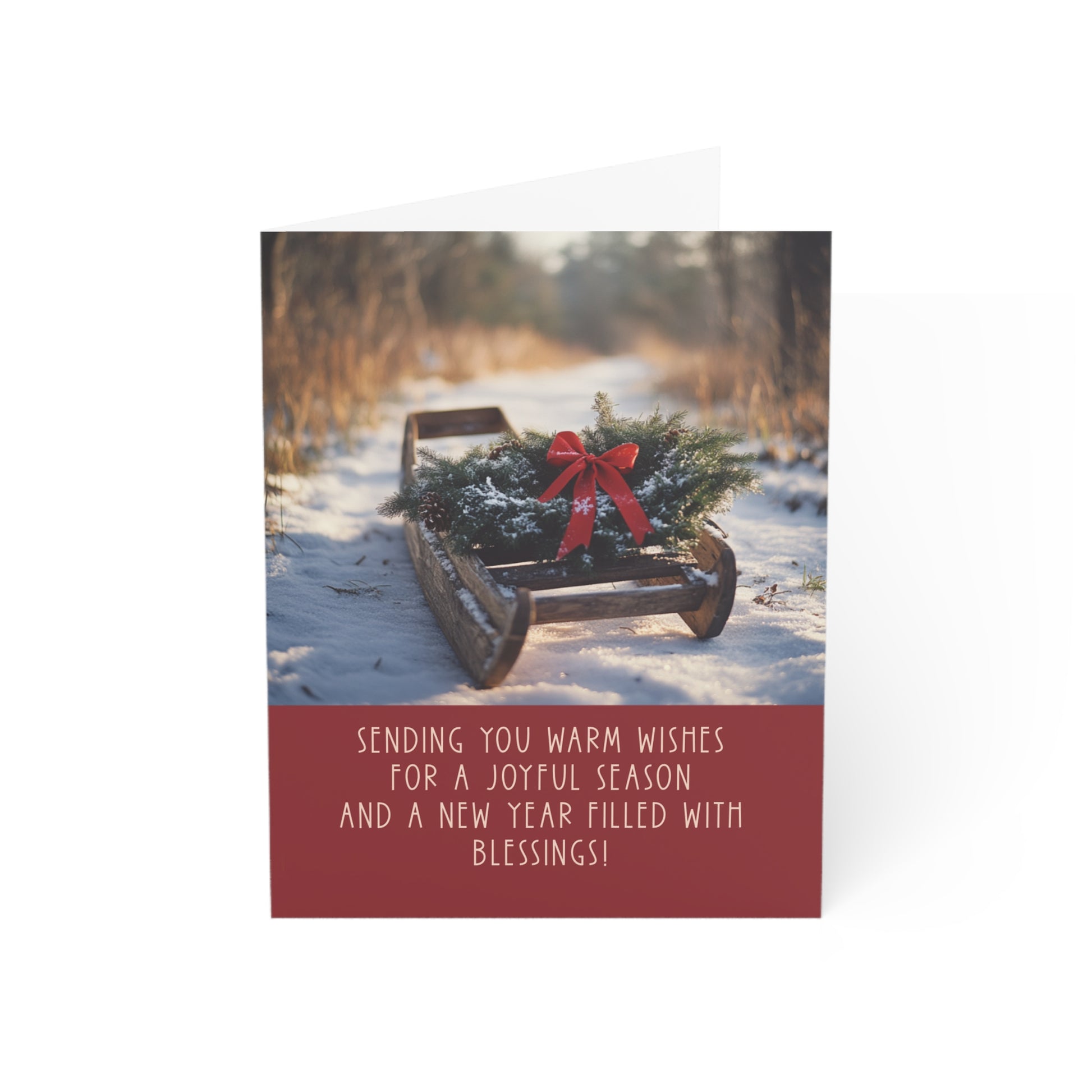 Holiday greeting card featuring a photograph of a vintage wooden sled resting on a snowy path. An evergreen wreath with a red bow adorns the sled. The card includes the message: 'Sending you warm wishes for a joyful season and a new year filled with blessings