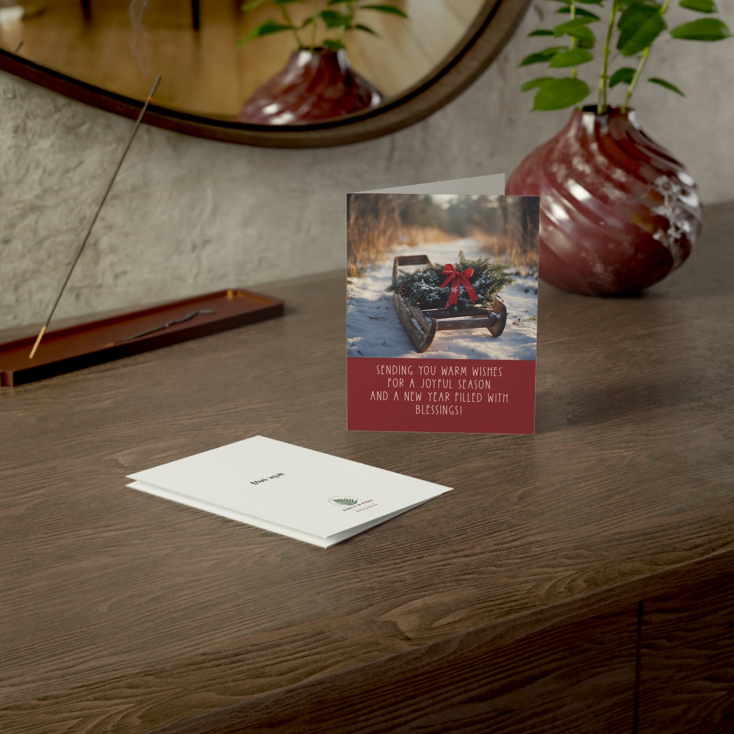 Mockup of holiday greeting card featuring a vintage wooden sled with an evergreen wreath and red bow on a snowy path. The front includes the message: 'Sending you warm wishes for a joyful season and a new year filled with blessings.' The inside is blank for a personalized message. The back features the Earth & Fern logo with the words 'Blank Inside‘.