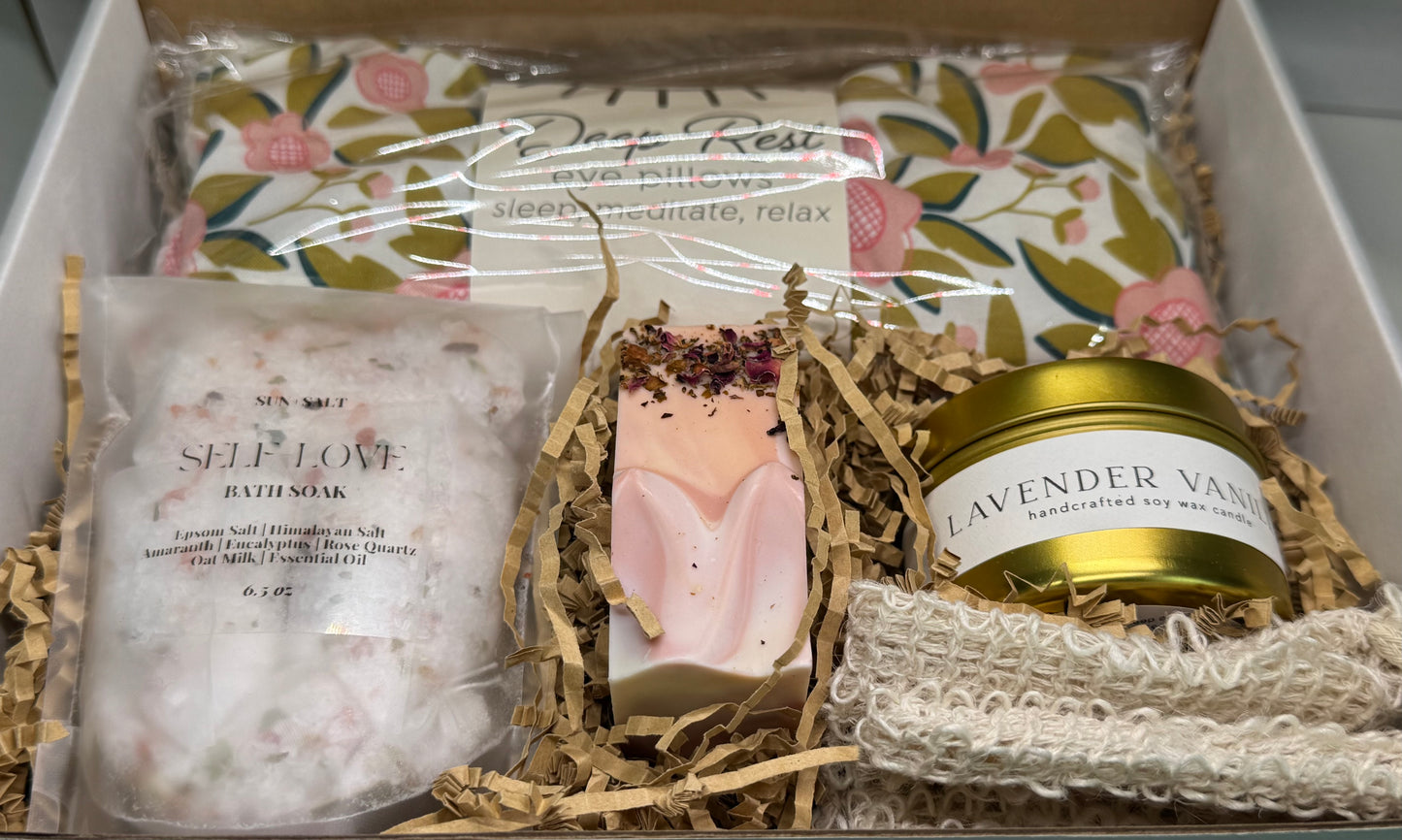 Self-care wellness gift box with lavender vanilla candle, handcrafted lavender rose soap, bath tea soak, and warm-cold therapie face pillow for relaxation and stress relief.

